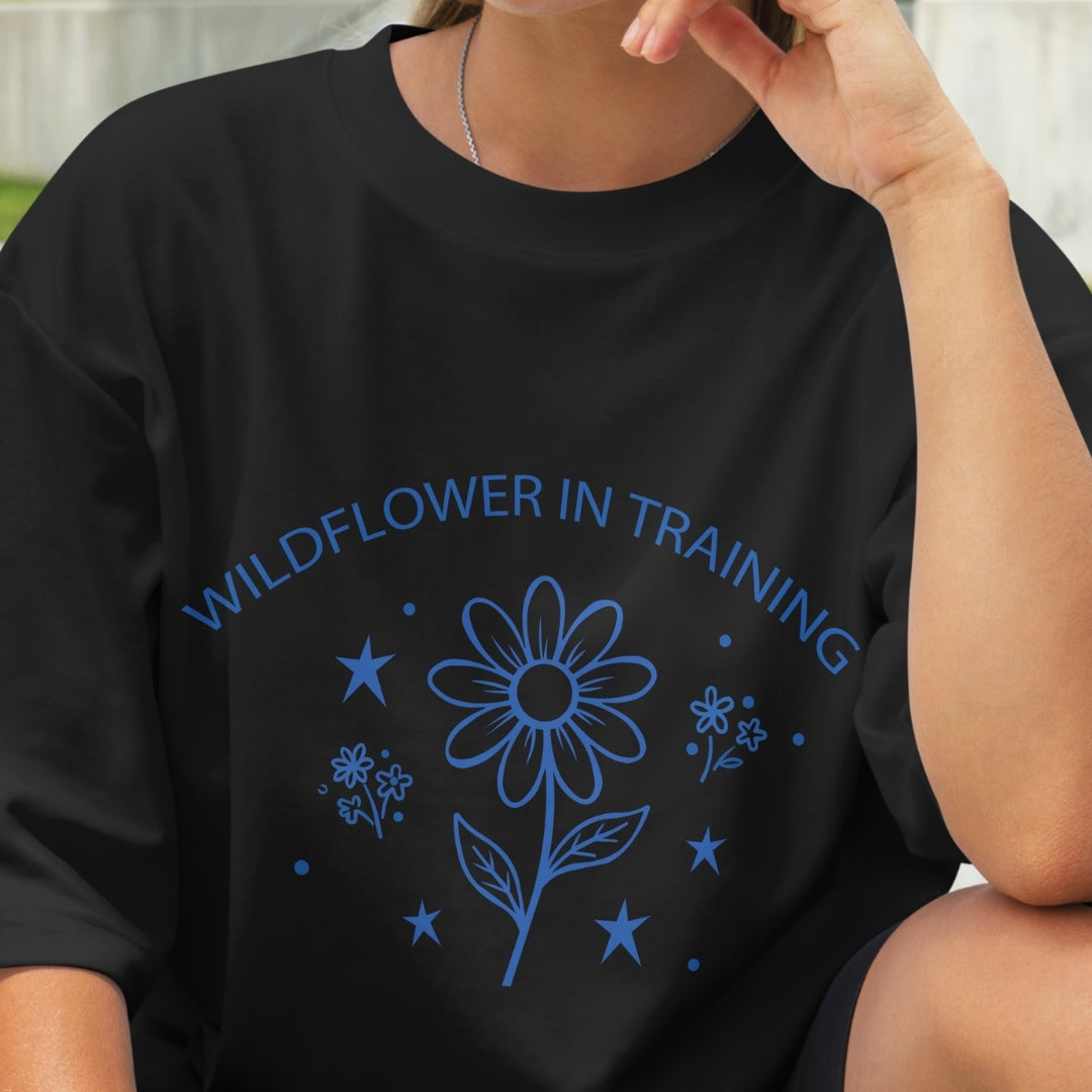 Black t-shirt with blue floral design and text 'Wildflower in Training' worn by a person.