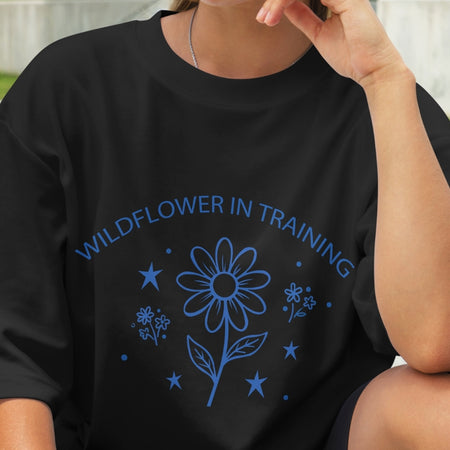 Black t-shirt with blue floral design and text 'Wildflower in Training' worn by a person.