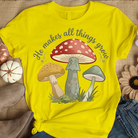 Yellow t-shirt with mushroom design and text 'He makes all things grow' on a textured surface.